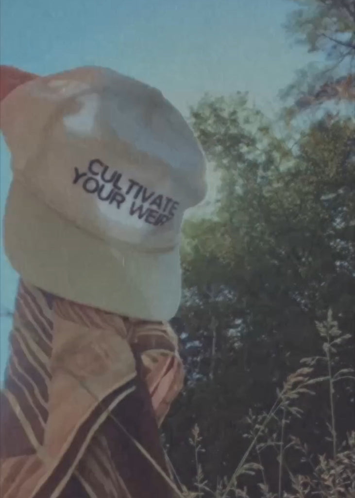 Person carrying a white cap with 'CULTIVATE YOUR WEIRD' text outdoors.