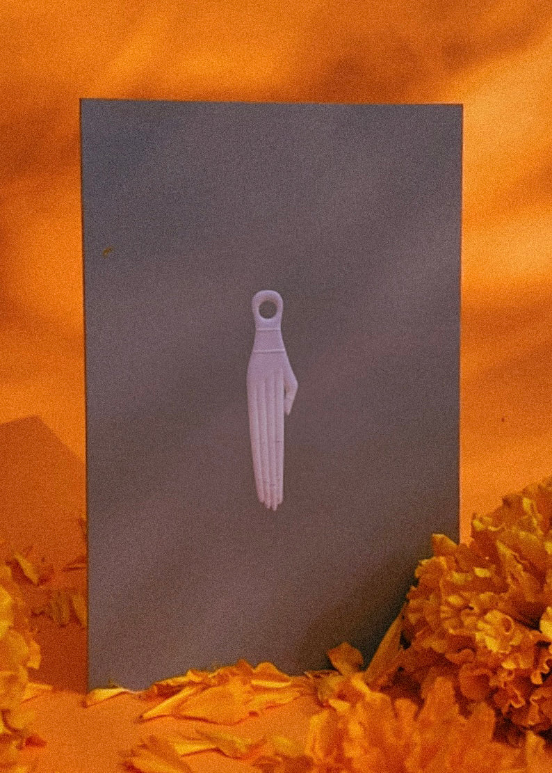 White khamsa hand amulet on a medium gray card with an orange textured background and marigolds
