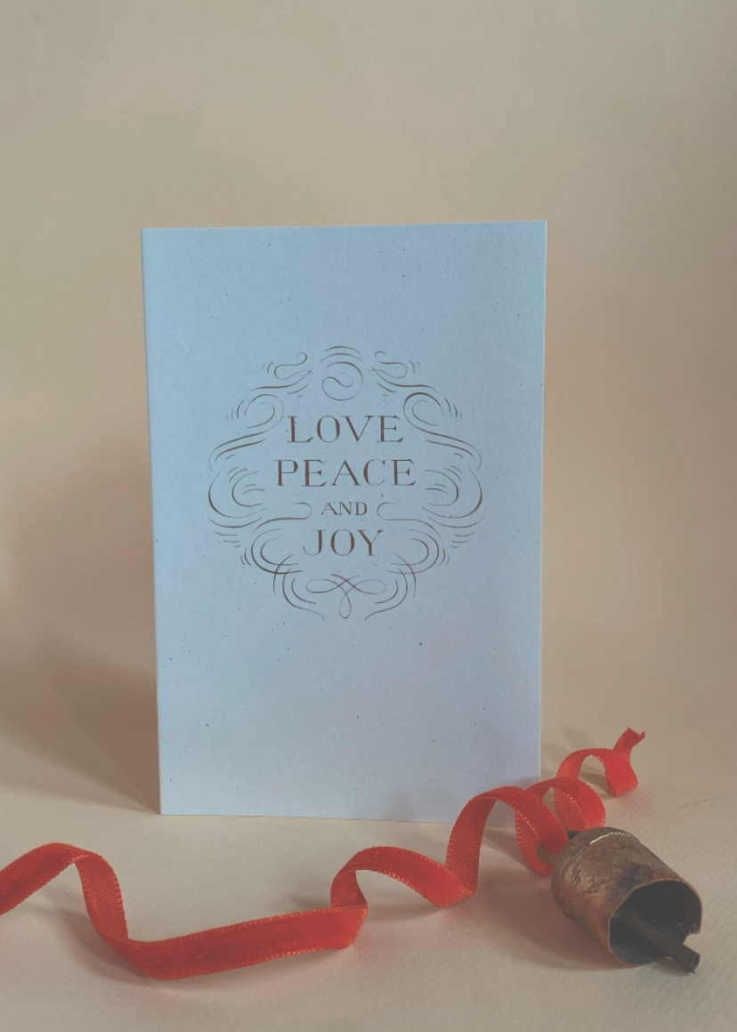 Ivory greeting card with 'Love Peace Joy' text on a beige surface with a red ribbon and rustic christmas bell.