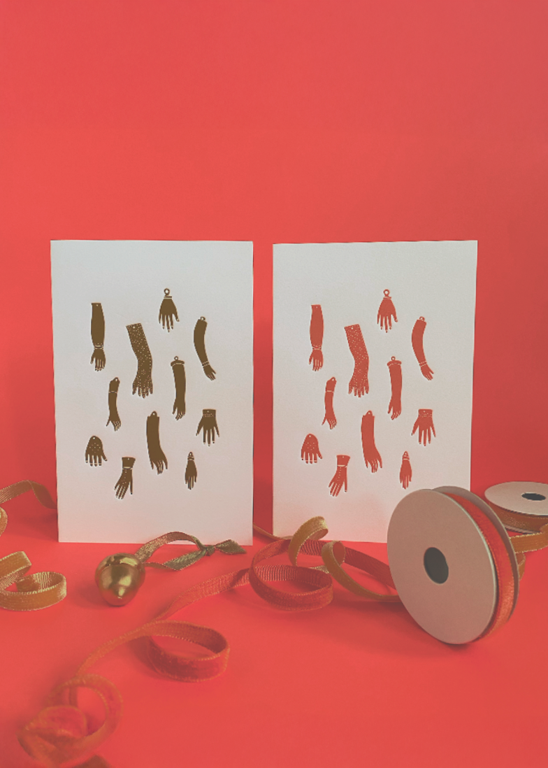 Two white cards with golden yellow and red doll arm and hand designs on a red background, accompanied by holiday ribbons and a golden christmas ornament