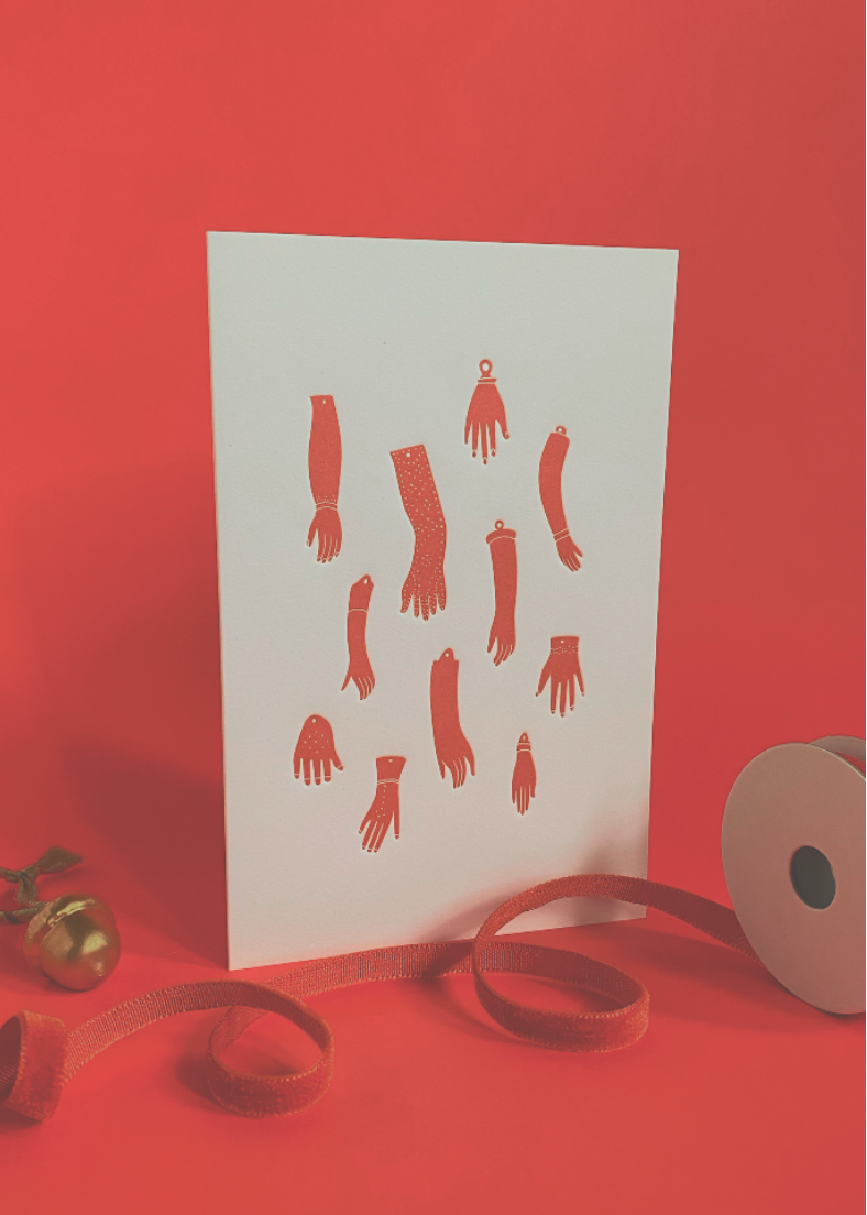 White card with rose colored doll arm shapes on a red background with ribbons and bells.